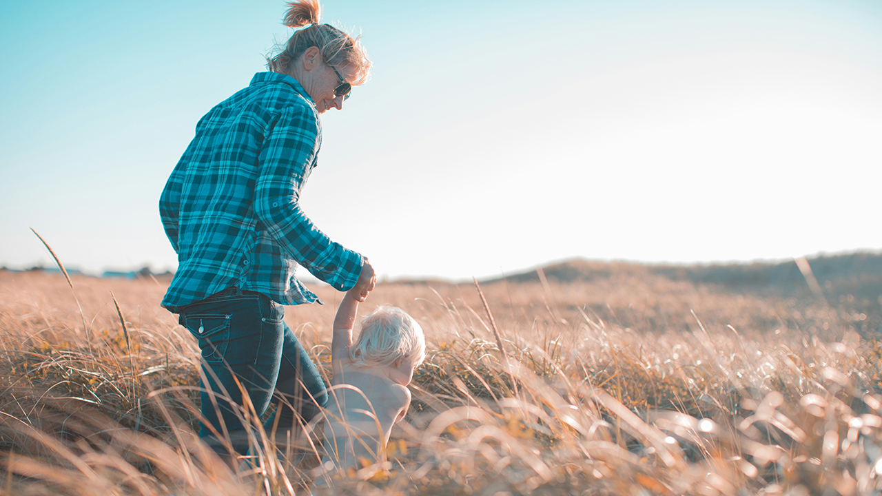 Mother walking hand-in-hand with her young child through a sunlit grassy field, evoking a warm, natural, and nurturing Mother’s Day moment.