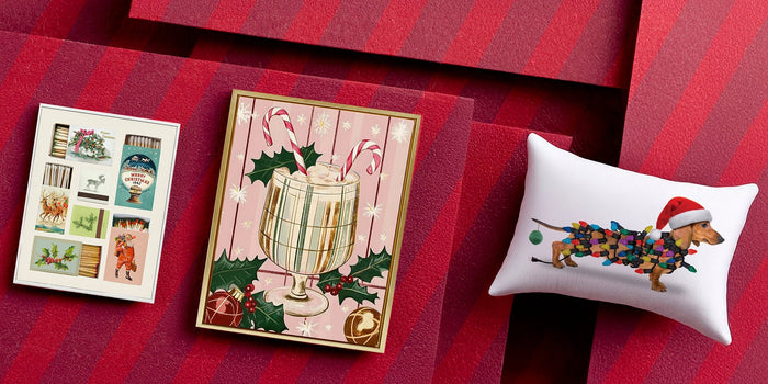 A holiday-themed flat lay featuring a vintage-style matchbook print, a framed illustration of a festive drink with candy canes and holly, and a decorative pillow with a dachshund wrapped in Christmas lights wearing a Santa hat, all arranged on red layers.